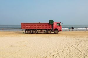 Red dump truck parked on a sandy beach with ocean waves and people in view.