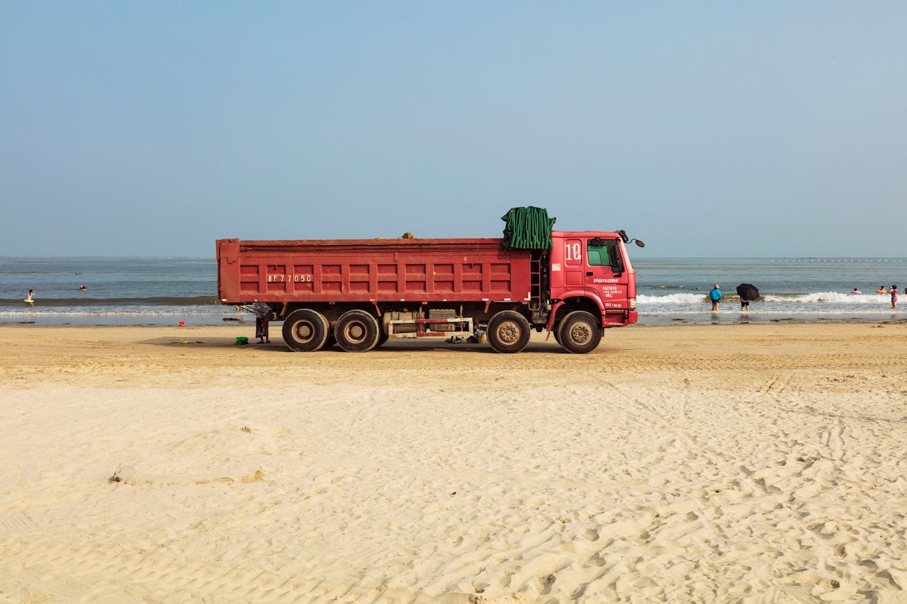 Red dump truck parked on a sandy beach with ocean waves and people in view.